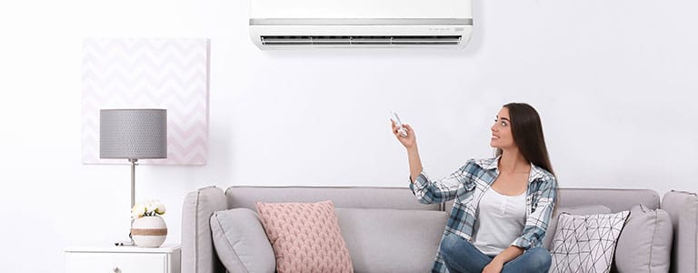 Ductless Systems Female homeowner sits on couch and uses remote to activate ductless system.