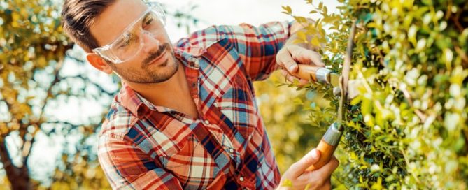 Man wearing safety glasses trimming hedges in from of Air Conditioning unit to prevent issues.